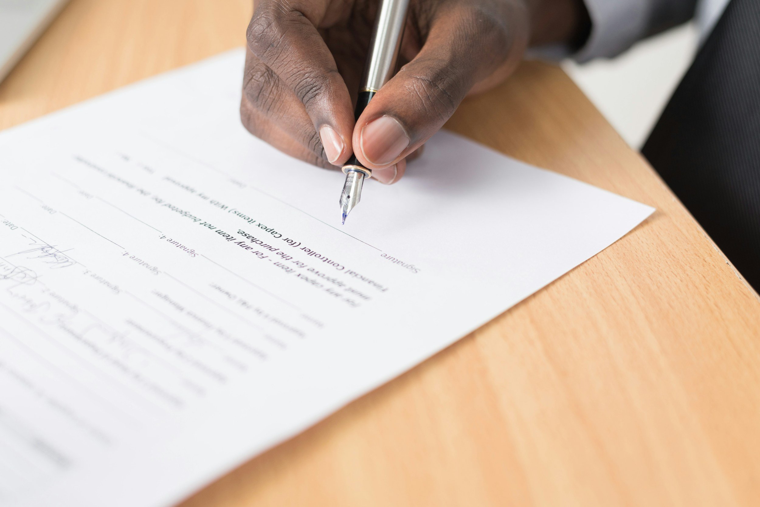 Person signing a document with a pen on a wooden desk.