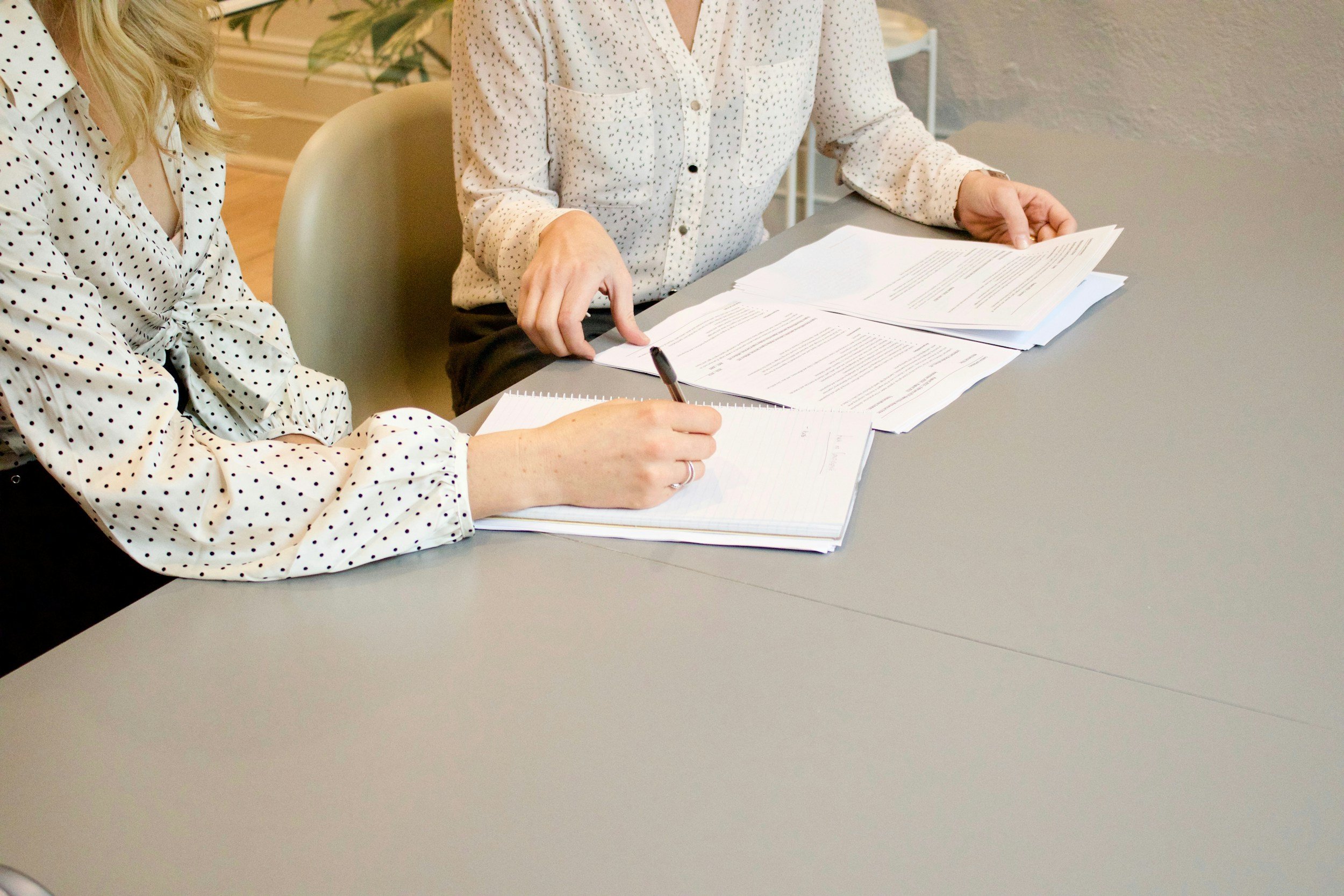 Two women sitting at a gray table with papers, one writing in a notebook and the other pointing at documents.