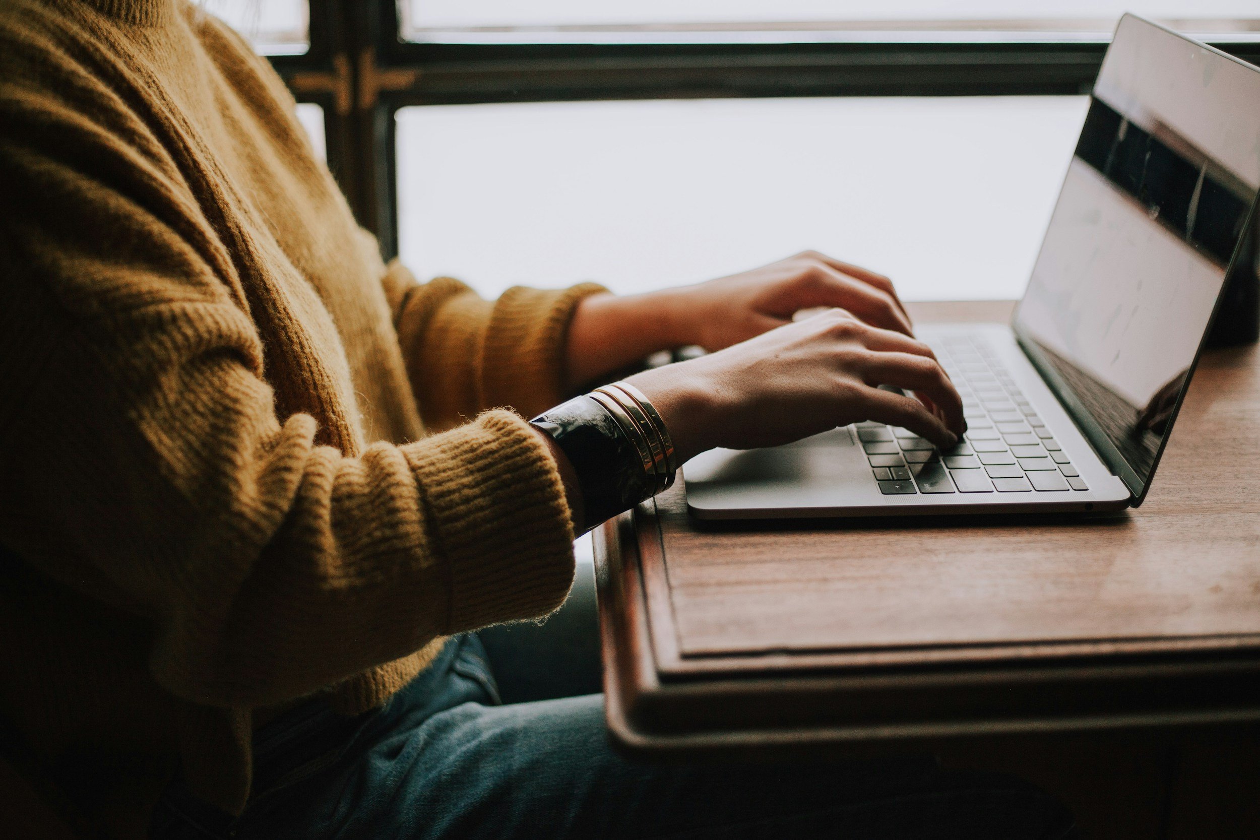 Person in a brown sweater using a laptop at a wooden table near a window.