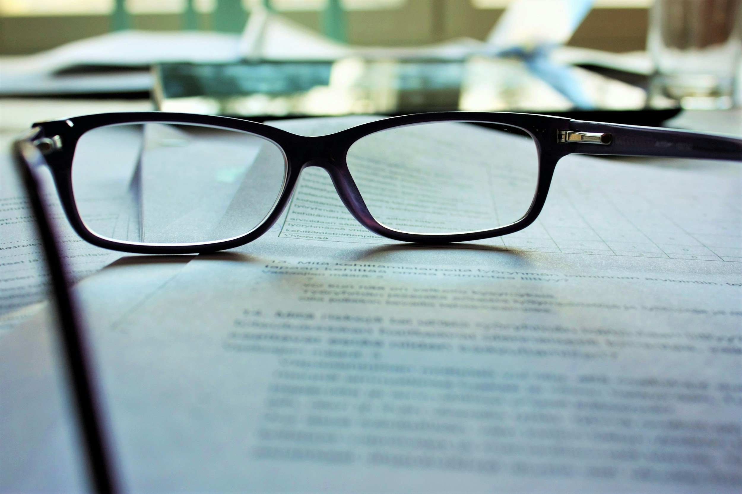 A pair of black reading glasses rests on an open newspaper on a table, with another pair of glasses blurred in the background.