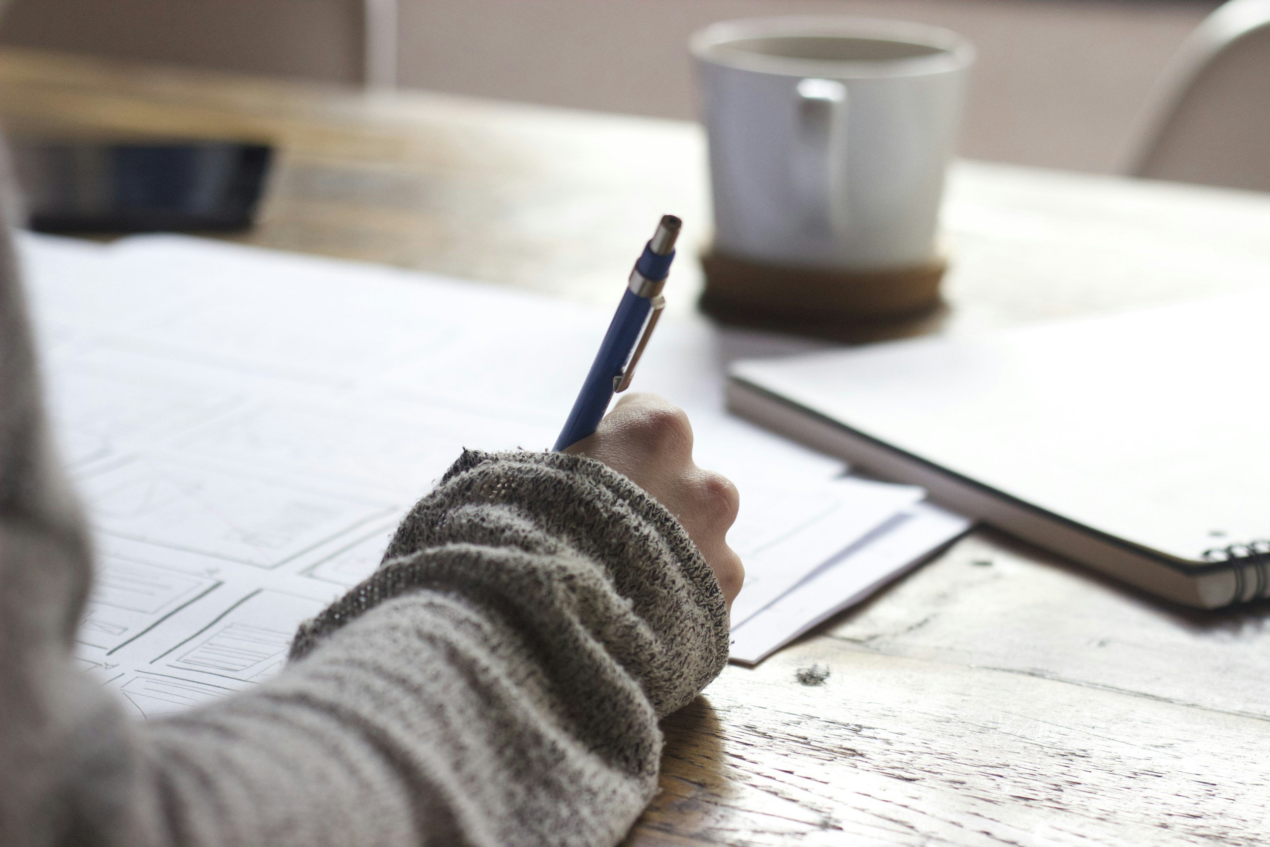 Person writing with a pen on papers on a wooden table, with a notebook and a coffee mug in the background.
