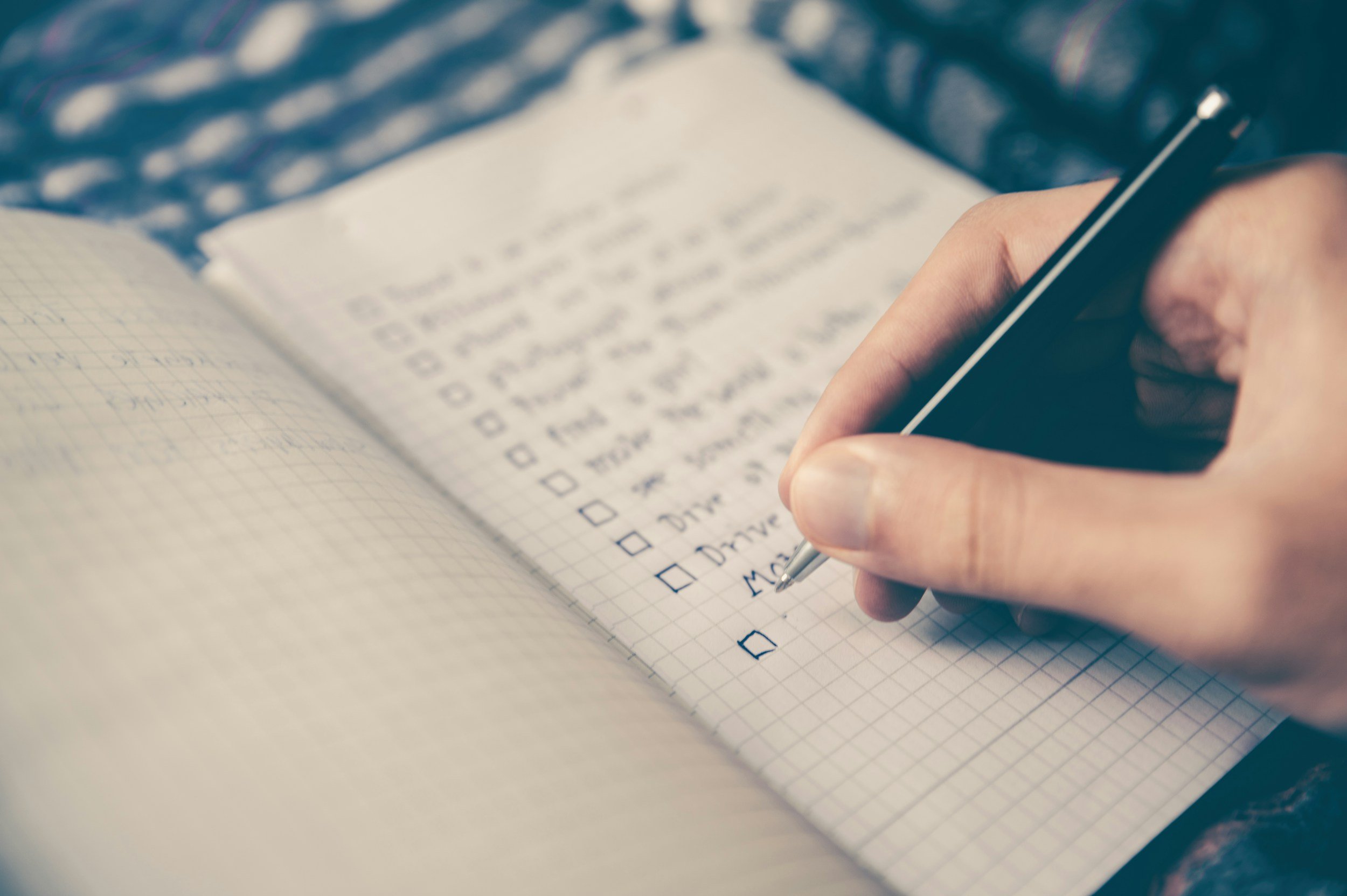 A person is writing a checklist in a graph paper notebook with a black pen, with a blurred background of a patterned blanket.