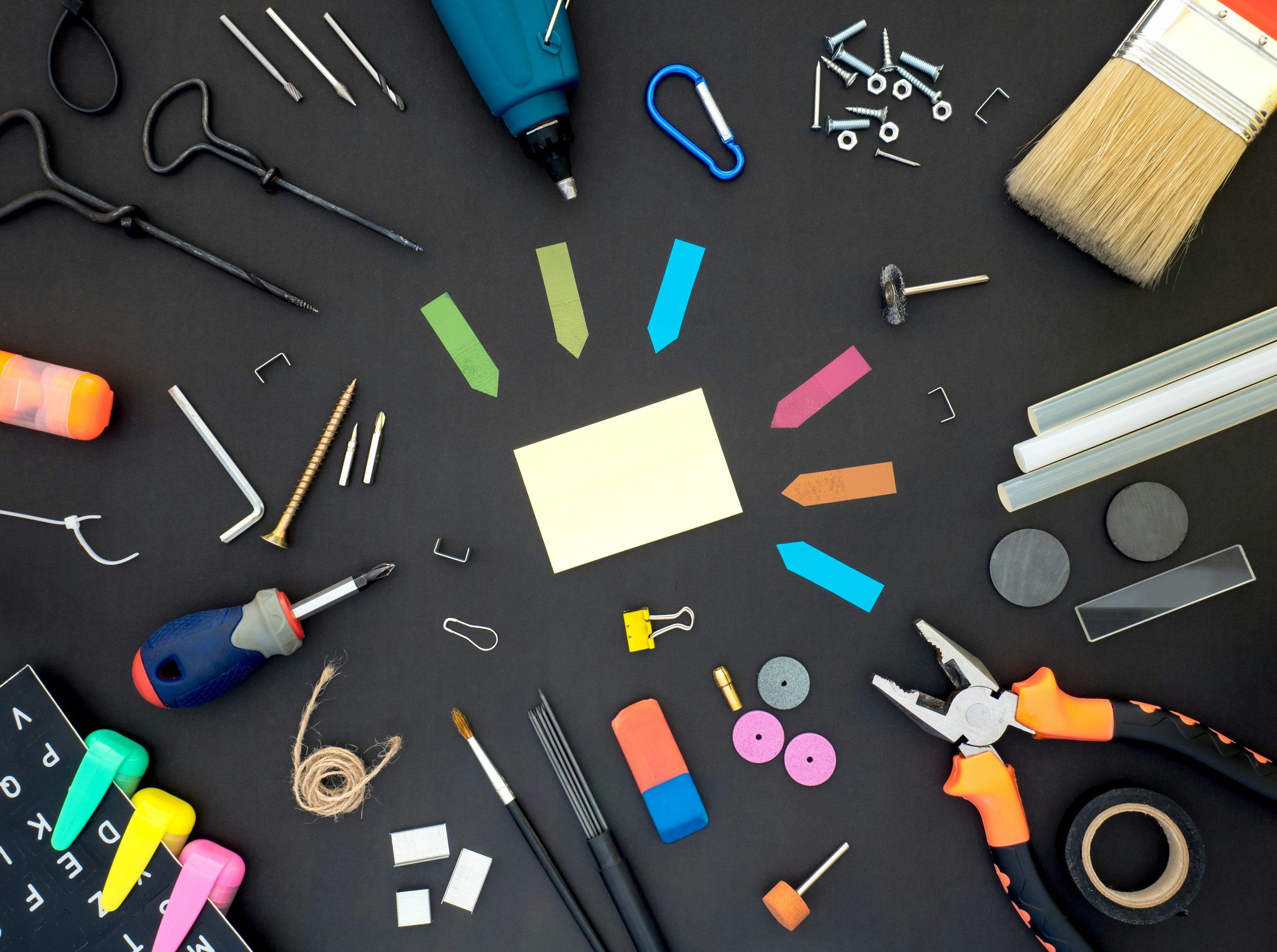 Assorted arts and crafts tools and supplies on black surface, including a paintbrush, scissors, yellow binder clip, glue sticks, various colored markers, thumbtacks, a drill, screws, nuts, bolts, a wire cutter, and colorful paper and tape.