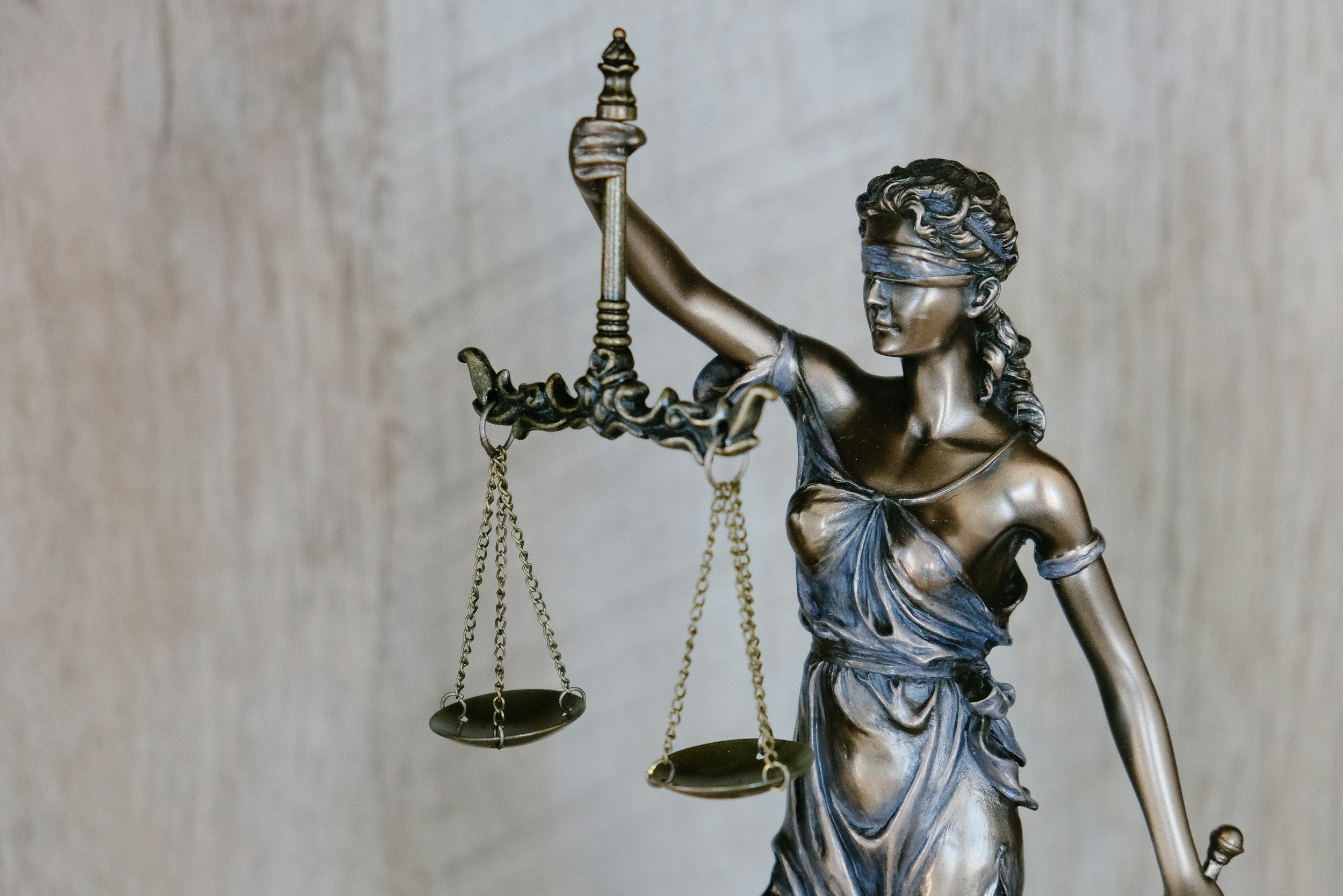 Bronze statue of Lady Justice holding a balance scale in her right hand, with a blindfold covering her eyes, standing against a neutral background.