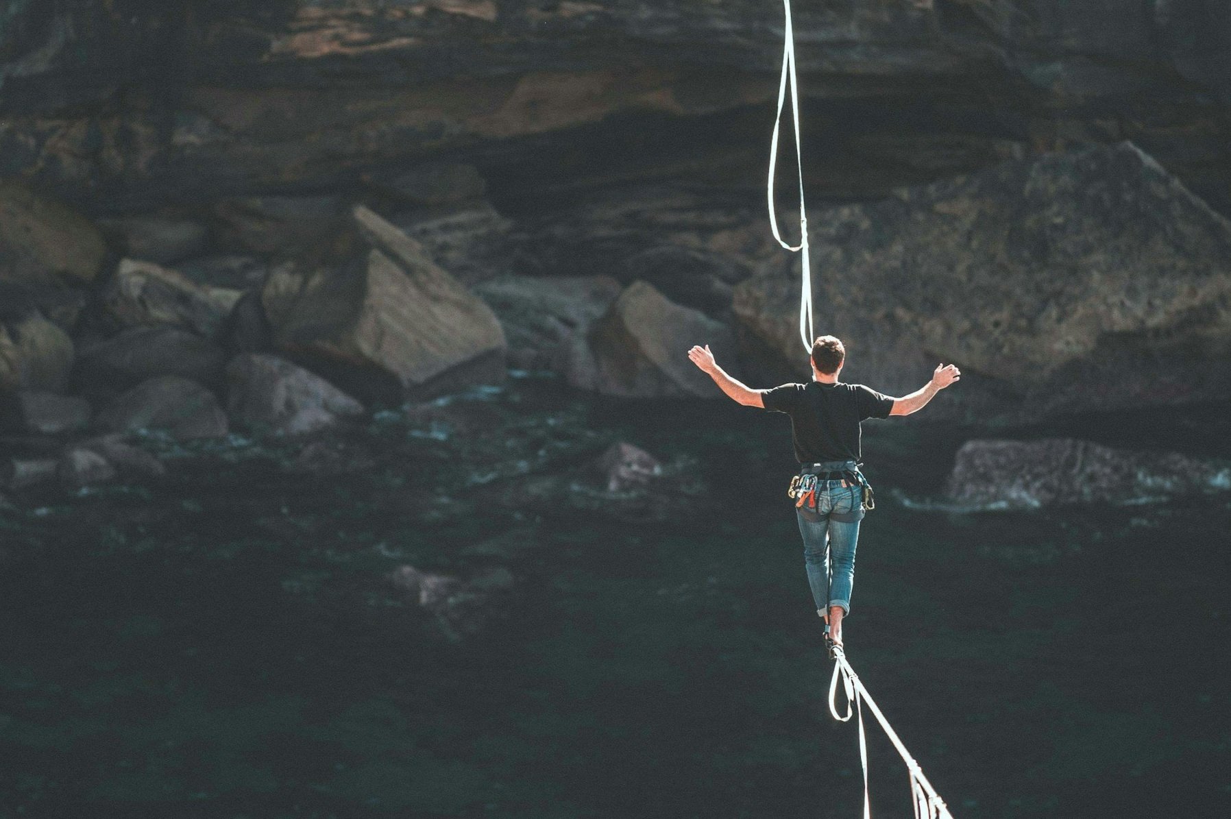 A person walking on a rope bridge over a body of water with large rocks and cliffs in the background.