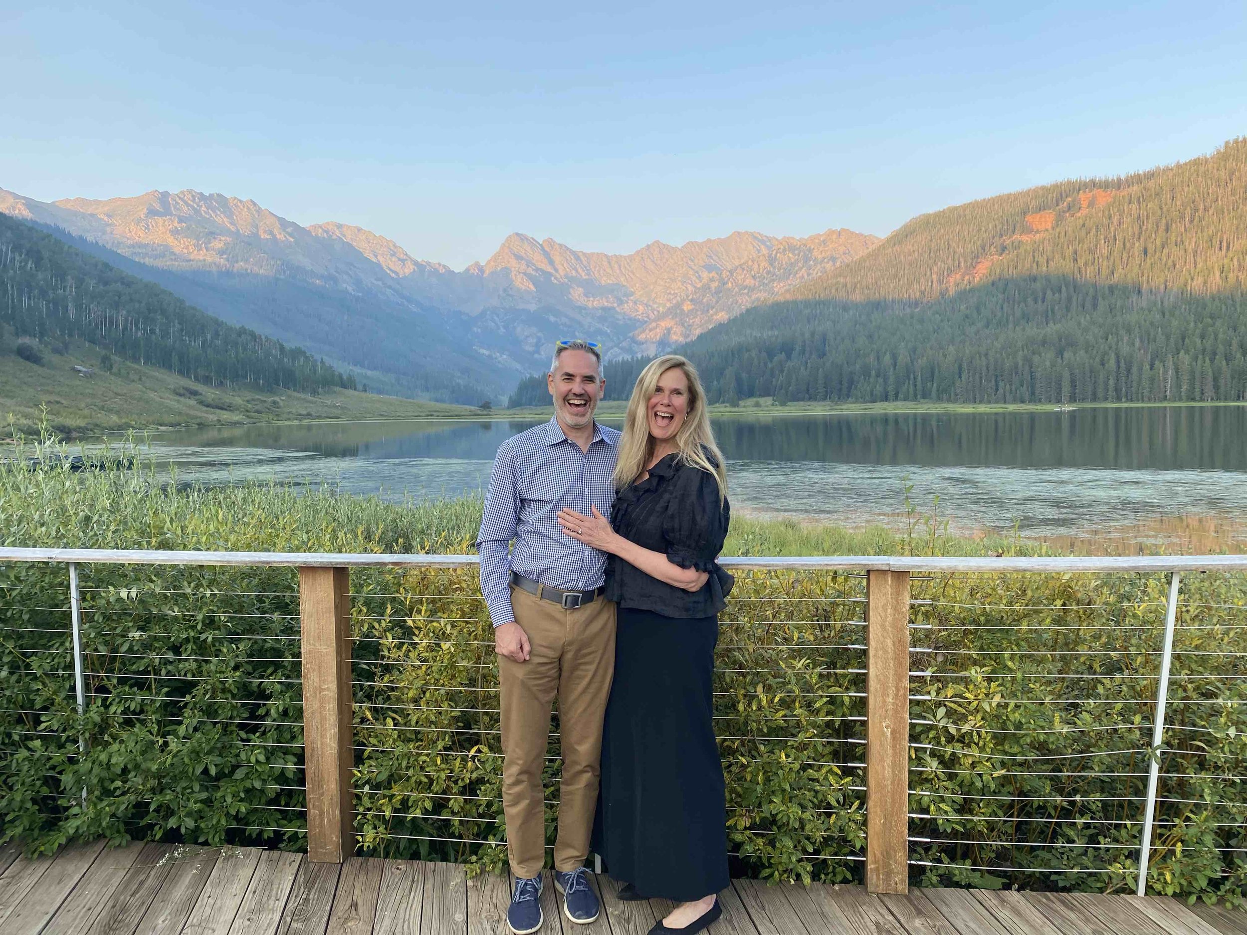 Darryl and Jeanie Shaper - A man and woman standing on a wooden deck with a lake and mountains in the background, smiling and posing for a photo.